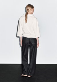 Bílá volná blůza se širokými rukávy, v kombinaci s tmavými džínovými kalhotami s širokými nohavicemi a oranžovým prošíváním. Pohled zezadu, minimální detaily.