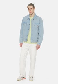 Ljusblå denimjacka med två bröstfickor, knappstängning, bärs över en ljusgrön skjorta, i kombination med vita byxor och grå sneakers.