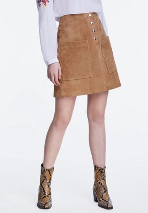 Vrouw draagt een beige suède A-lijn rok met knopen en grote voorzakken, gecombineerd met enkellaarsjes met slangenprint en een wit bloesje.