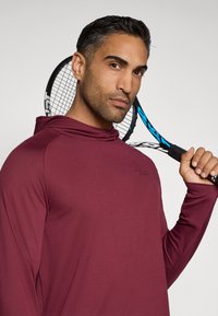 Maroon long-sleeve hoodie, featuring a relaxed fit and a subtle logo. The model holds a tennis racquet against a light background.