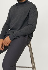 Mann sitzt auf einem Metallhocker, trägt einen dunkelgrauen Pullover mit langen Ärmeln und dunkle Hosen, die Hände ruhen im Schoß vor einem weißen Hintergrund.