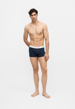 Mladi muškarac mišićave građe, obučen u tamnoplave Tommy Hilfiger bokserice, stoji bos na jednostavnoj bijeloj pozadini.