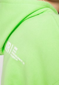 Felpa con cappuccio verde neon con una texture liscia, caratterizzata da un logo bianco stampato sul davanti e un cappuccio con coulisse.