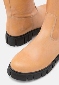 Botas de tornozelo em couro castanho com um acabamento mate, solas de borracha preta em contraste e detalhes de costura sutis. Logotipo da marca estampado na lateral.