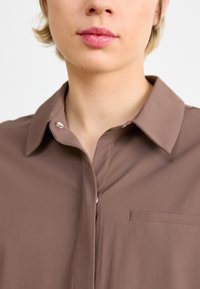 Camicia color marrone con un tessuto liscio, dotata di una piccola tasca sul petto, chiusure con bottoni a pressione e dettagli di cucitura pulita.