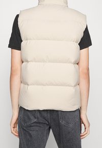 Person som bär en beige vadderad ärmlös väst över en svart kortärmad skjorta och mörkgrå jeans, sedd bakifrån.