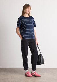 Marineblå stripete t-skjorte med korte ermer, svarte tapered bukser og rosa sneakers med hvite detaljer. Modellen holder en svart tote-bag. Enkelt design og farger.
