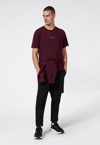 Man in een bordeaux Champion t-shirt met een bijpassende sweatshirt om de taille gebonden, zwarte broek en zwarte sneakers tegen een witte achtergrond.