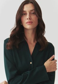 Camicia button-down verde scuro con maniche lunghe, realizzata in tessuto liscio. Presenta una leggera lucentezza e un design sartoriale. Dettagli in bottone dorato visibili.
