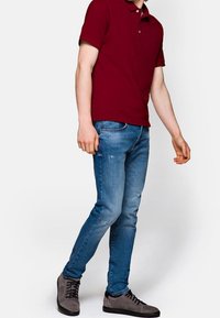 Burgundské polo tričko, světlé modré úzké džíny s odřenými detaily a šedé semišové tenisky s černými akcenty, model stojí na neutrálním pozadí.