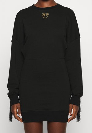 Personne portant une robe noire à manches longues avec un emblème doré près de l'encolure et des poignets de manches à franges.