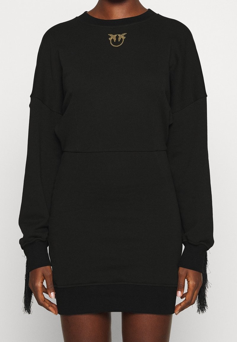 Personne portant une robe noire à manches longues avec un emblème doré près de l'encolure et des poignets de manches à franges.