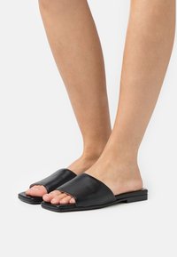 Sandales à talon plat en cuir noir avec un design supérieur courbé, bout ouvert et semelle plate. Texture lisse avec un style minimaliste.