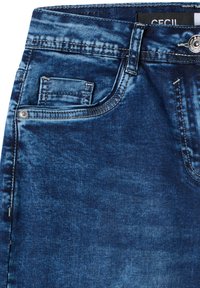 Gros plan d’un jean en denim bleu acidulé montrant la poche avant, la boucle de ceinture, le bouton, et une partie de la ceinture avec une étiquette à l’intérieur.