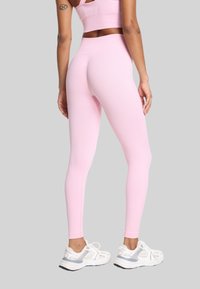 Kobieta w różowych legginsach z wysokim stanem i pasującym topie crop, w białych sneakerach, stojąca na tle szaro-skalnym.