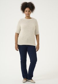 Pull en maille léger beige, à manches courtes avec un motif texturé ; associé à un jean droit bleu foncé et des ballerines de couleur claire.
