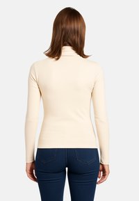 Maglione beige a collo alto con maniche lunghe, realizzato in un tessuto liscio. Indossato con jeans attillati blu scuro, che mostrano una vista posteriore pulita.
