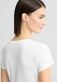 T-shirt blanc à manches courtes avec un col rond, texture lisse et coupe décontractée, présenté sur une personne de dos.