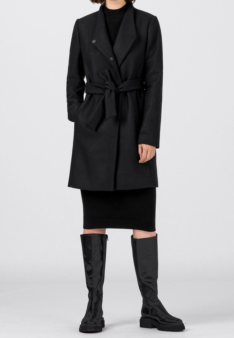 Musta vyöllinen takki, joka on valmistettu teksturoidusta kankaasta ja jossa on kaksirivinen malli sekä leveät kaulukset; yhdistetty polven korkeisiin kiiltäviin mustiin saappaisiin.