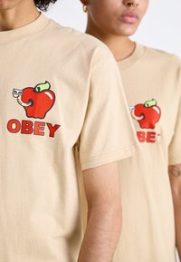 Beige bavlněná trička s grafikou červeného jablka se červem a slovem "OBEY" v tučných červených písmenech, kulatý výstřih, uvolněný střih.