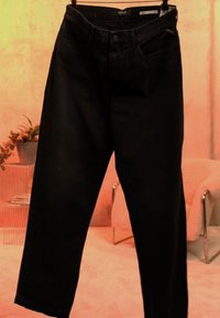 Sorte denimjeans hængende på en klemmebøjle i et blødt oplyst rum med en beige lænestol og en plante i baggrunden.