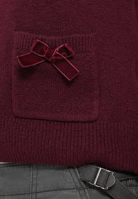 Maglione color vino con superficie testurizzata, caratterizzato da una piccola tasca con un dettaglio a fiocco di velluto. Orlo e polsini a coste.