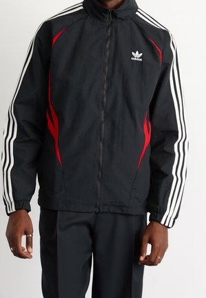 Mężczyzna nosi czarną kurtkę sportową Adidas z białymi paskami na rękawach i czerwonymi akcentami, zapinaną na zamek, ręce swobodnie opuszczone wzdłuż ciała.