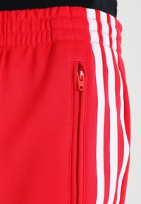 Červené sportovní kalhoty s texturovaným materiálem, které mají bílý pruhový design a boční kapsu na zip. Elastický pas pro bezpečné nošení.