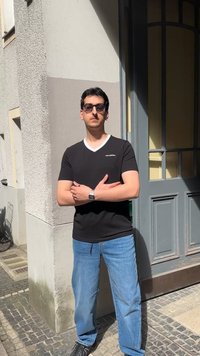 Άντρας με γυαλιά ηλίου, μαύρο μπλουζάκι, μπλε τζιν, στέκεται με σταυρωμένα χέρια έξω κοντά σε μια πόρτα μια ηλιόλουστη μέρα.