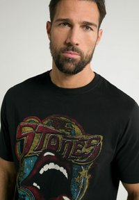 Schwarzes T-Shirt aus Baumwolle mit einem bunten grafischen Druck eines Mundes und Sternen, mit dem Text "The Stones" in stilisierten Buchstaben.