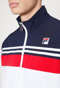 Track jacket in navy, red, and white with horizontal stripes. Features a zip closure and logo tag. Made from smooth, lightweight fabric.