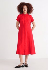 Κόκκινο φόρεμα με κουμπιά και κοντά μανίκια, μέχρι το γόνατο, φτιαγμένο από ελαφρύ ύφασμα. Συνδυάζεται με μαύρες σαγιονάρες με λουριά, λεία υφή.