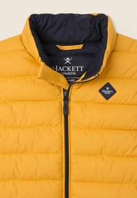Żółta ocieplana kurtka z granatowym podszewką, z poziomym przeszyciem, czarnym zamkiem błyskawicznym i łatą z diamentowym logo po lewej stronie.