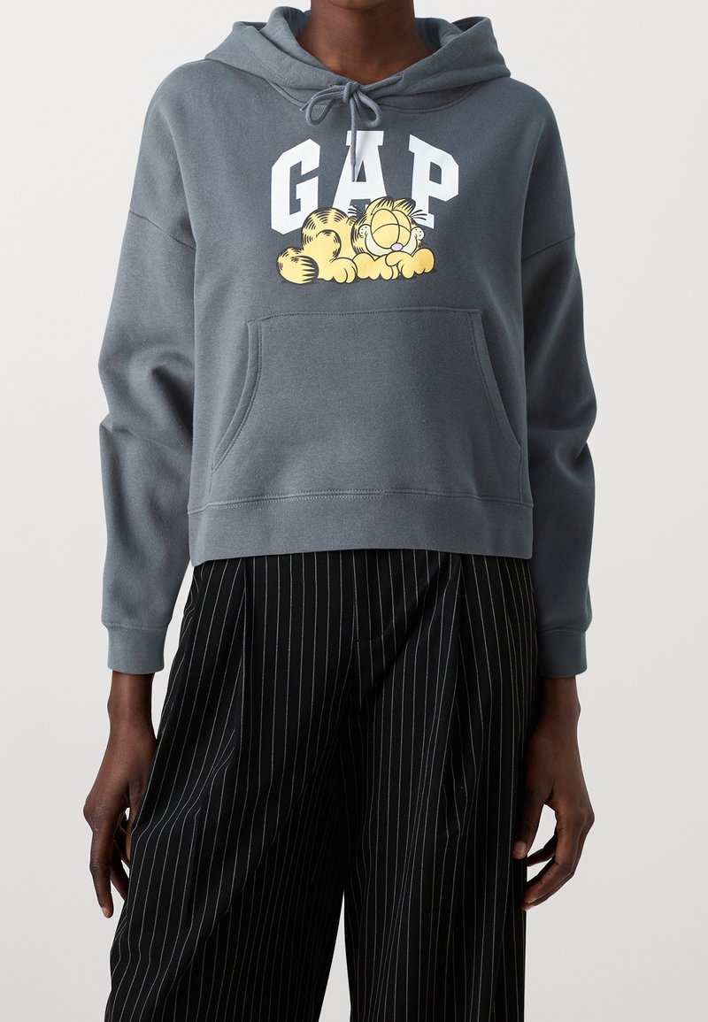 Felpa corta grigia con tasca frontale, caratterizzata da un logo bianco "GAP" e da un design di un gatto cartoon giallo. Il materiale sembra morbido e testurizzato.