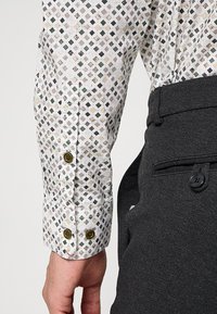 Het shirt met lange mouwen is gemaakt van witte stof met een geometrisch patroon in groen, zwart en beige. Er zijn donkergrijze broeken zichtbaar met een knoopdetail.