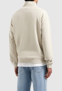 Beige sweatshirt met een hoge kraag en geribbelde manchetten, met een ontspannen pasvorm. Draag het over een wit shirt, gecombineerd met lichtblauwe jeans.