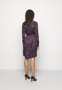 Lila och svart mönstrad klänning med lång ärm, ett bälte i midjan och knälång fåll, kombinerad med svarta stilettklackar.