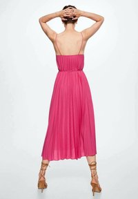 Robe fuchsia plissée avec un corsage ajusté et de fines bretelles, arrivant mi-mollet. Portée avec des talons marron à fines brides, vue de dos.