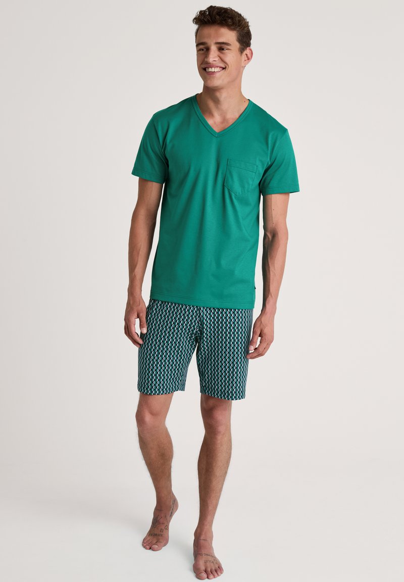 Groene katoenen V-hals T-shirt met een zak, gecombineerd met gestreepte blauwe en witte shorts. Model staat blootvoets en lacht tegen een neutrale achtergrond.