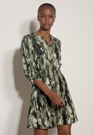 Femme portant une robe à manches longues à motifs verts et noirs, boutonnée à l'avant avec une taille cintrée, debout devant un fond clair uni.