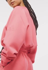 Ružová oversized mikina s úzkym pásom a textúrovanou látkou. Obsahuje široké rukávy a jemné logo blízko ramena.
