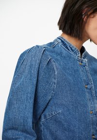 Denim jack met opgezette mouwen, een staande kraag, een sluiting met knopen aan de voorkant en twee zijzakken. De kleur is medium blauw met een gestructureerde afwerking.