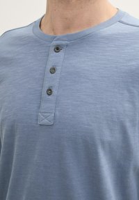 Világoskék henley póló, puha pamutból készült, kerek nyakkivágással, három gombos légbuborék-nyitással sötét gombokkal és texturált felülettel.