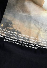 Czarny tekstyl z napisami o spokoju, z jasną grafiką morza, podkreślającą fale i wewnętrzną siłę.