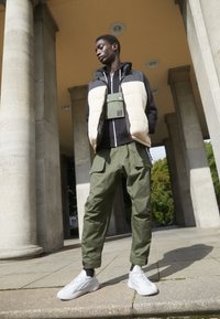 Väst i beige och svart över en svart hoodie, kombinerat med olivgröna cargobyxor och vita sneakers, framför stenpelare utomhus.