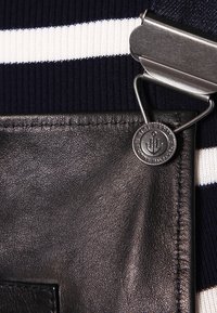 Zwarte leren patch met zilveren hardware en een cirkelvormig logo, tegen een marineblauwe en witte gestreepte stof. Textuurdetails zijn zichtbaar.