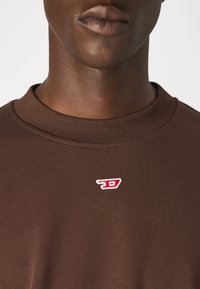 Felpa marrone a maniche lunghe con colletto a girocollo, caratterizzata da un piccolo logo ricamato rosso e bianco sulla scollatura. Texture morbida, design aderente.