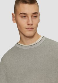 Szary sweter robiony na drutach z okrągłym dekoltem, z delikatną teksturą i kontrastowym białym wykończeniem wewnętrznego kołnierza. Zbliżenie na szczegóły materiału.