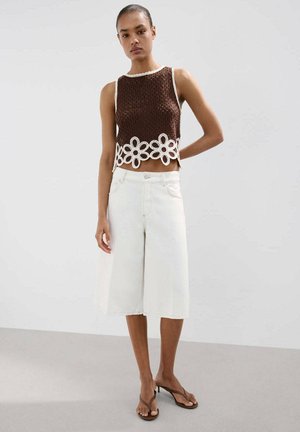 Femme portant un haut en crochet marron sans manches avec bordure florale blanche, pantalon blanc large et court, et sandales marron à talons, debout immobile.