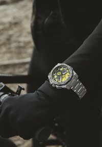 Sudraba nerūsējošā tērauda pulkstenis ar dzeltenu un melnu ciparnīcu, kas aprīkots ar hronogrāfu, izteiksmīgiem atzīmējumiem un saiteņu aproci.